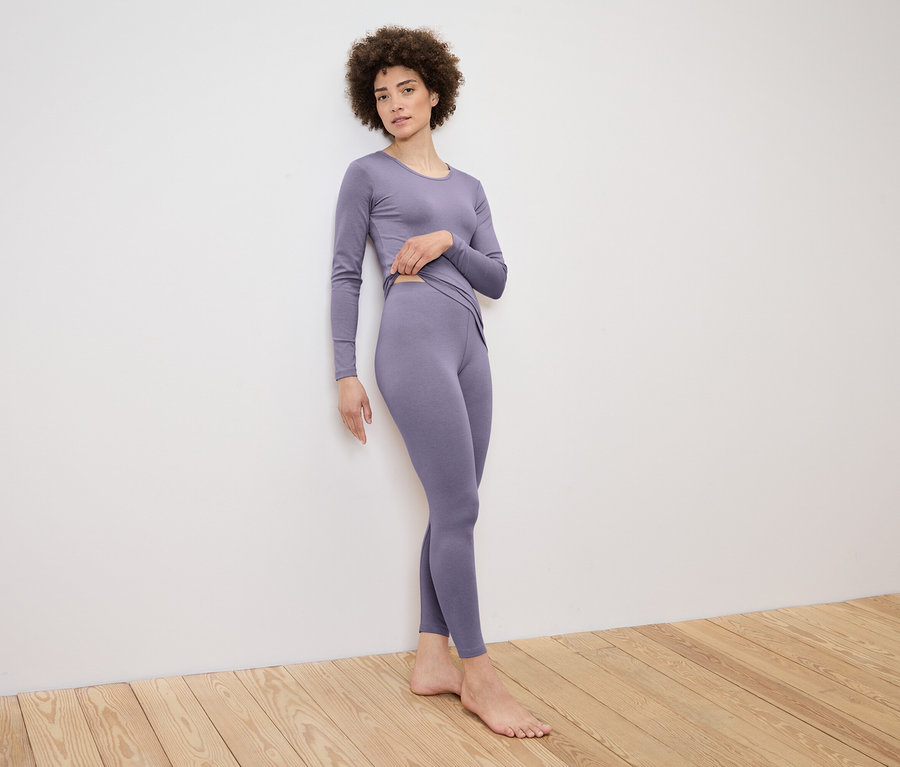 Frau posiert in einem lila farbenen Longsleeve-Top und Leggings-Set an einer Wand.