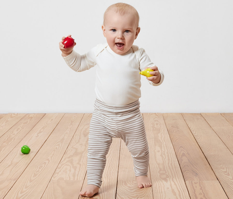 Ein lächelndes Baby steht auf einem Holzboden und hält ein rotes und ein gelbes Spielzeug in den Händen. Es trägt eine 5 Kinder-Leggings.