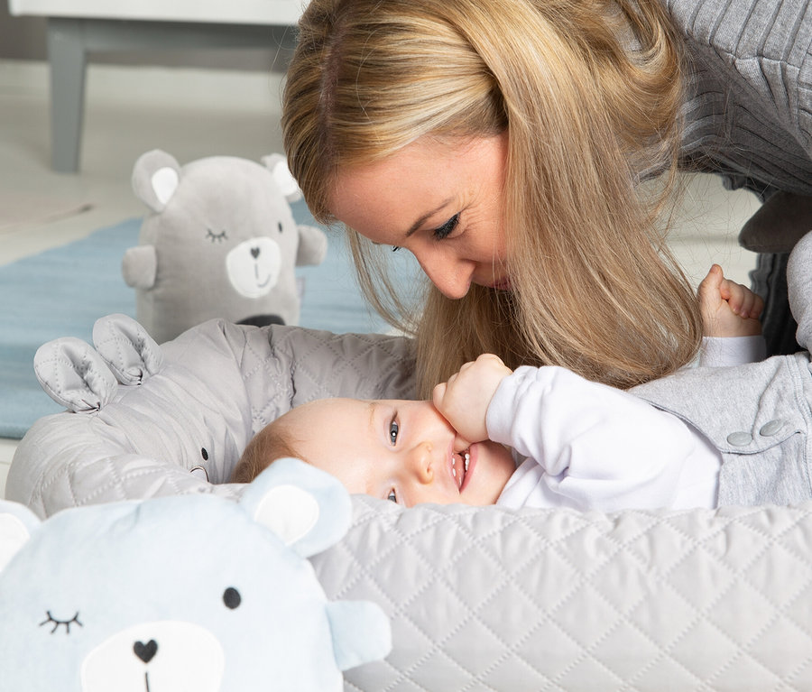 Mutter schaut auf ein lächelndes Baby, das in einer grauen Roba Babylounge liegt, umgeben von Plüschtieren.