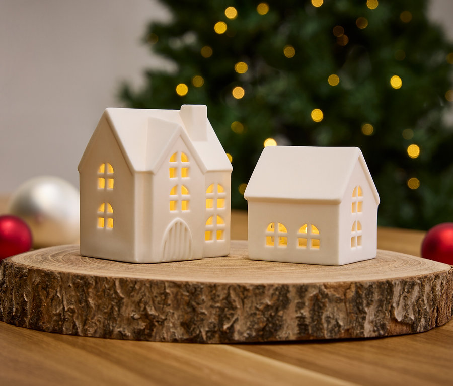 Zwei weiße Häuschen auf einem Holzsockel mit Weihnachtsschmuck und Baum im Hintergrund.