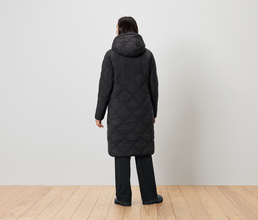 Eine Frau steht mit dem Rücken zur Kamera und trägt einen schwarzen Steppmantel mit Kapuze.