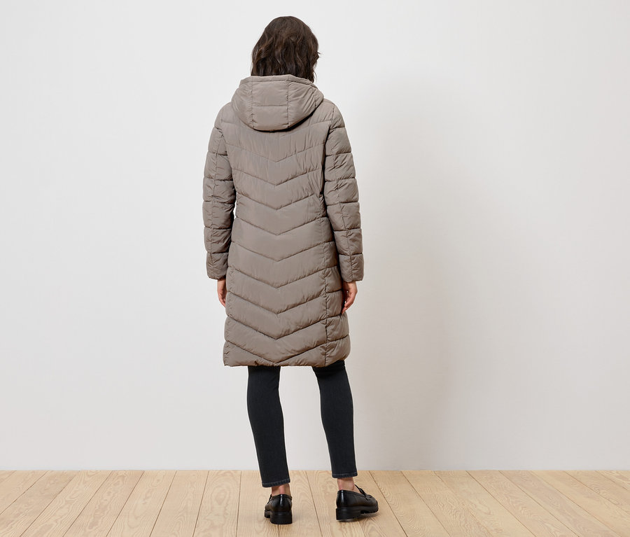 Frau von hinten trägt einen langen Steppmantel mit Kapuze, schwarze Hose und schwarze Schuhe.