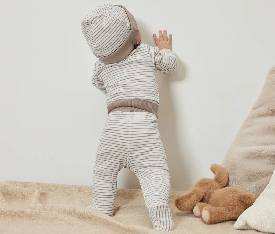 Ein Baby in einem 3-teiligen Baby-Set steht an einer Wand.