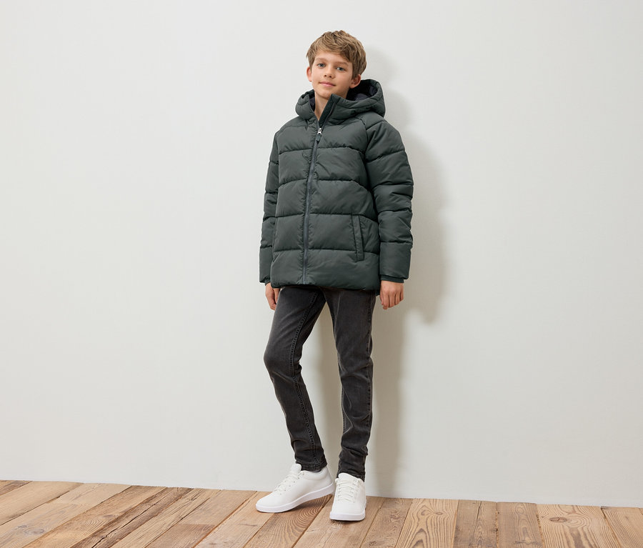 Ein Junge steht und trägt eine dunkelgrüne Daunenjacke, graue Jeans und weiße Turnschuhe.