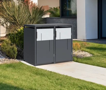 Ein grauer Mülltonnencontainer mit zwei Türen steht auf einem gepflasterten Weg neben einem Haus.