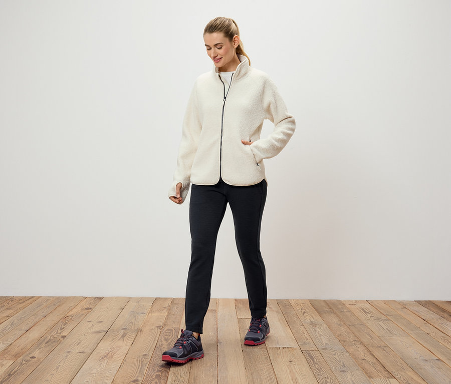 Eine Frau in einer cremefarbenen Fleecejacke, schwarzen Hosen und Sportschuhen steht auf einem Holzboden vor einer weißen Wand.