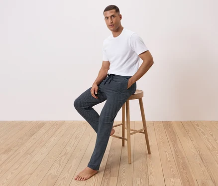 Mann sitzt auf einem Hocker, trägt ein weißes T-Shirt und gemusterte Hosen.