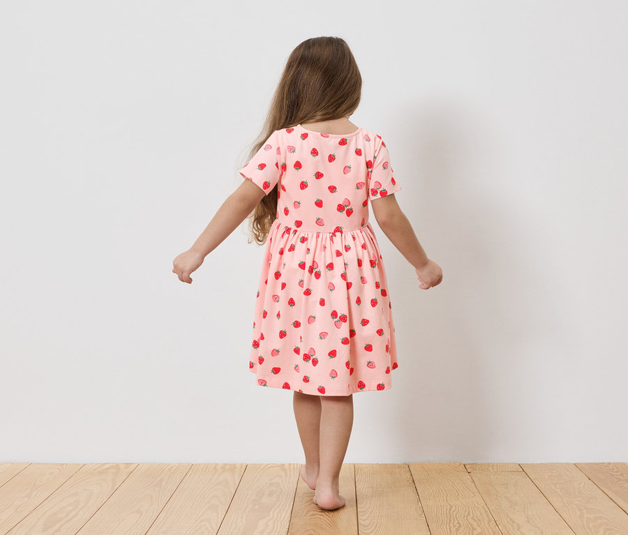 Mädchen von hinten in einem hellrosa Kleid mit Erdbeermuster, auf Holzboden stehend.
