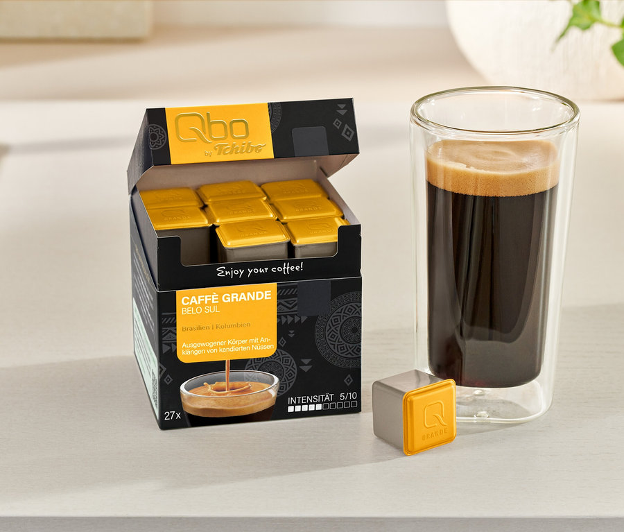 Eine geöffnete Packung Qbo Caffè Grande BELO SUL mit Kapseln, eine einzelne Kapsel und ein Glas Kaffee stehen auf einem Tisch.