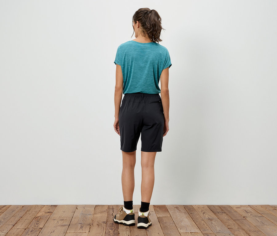 Rückansicht einer Frau mit Pferdeschwanz, die ein blaugrünes T-Shirt, schwarze Shorts und Turnschuhe auf einem Holzboden trägt.