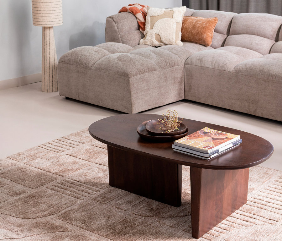 Ein Wohnzimmer mit einem beigen Ecksofa, einem dunkelbraunen ovalen Couchtisch mit Zeitschriften und einer Dekoschale, einer beigen Stehlampe und einem gemusterten Teppich.