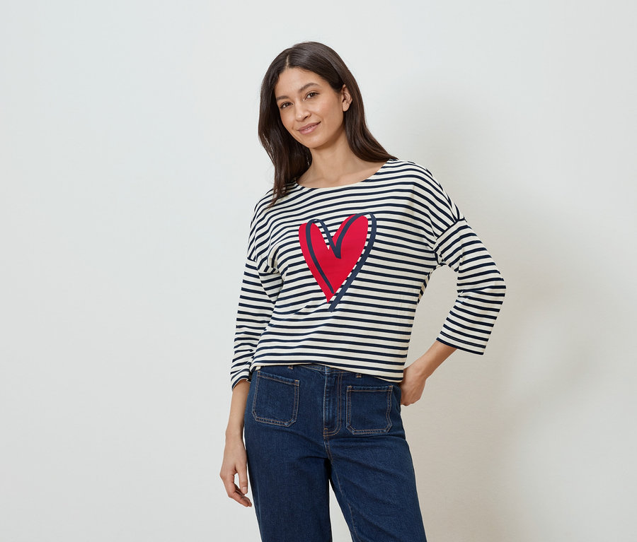 Frau trägt ein gestreiftes Langarmshirt mit einem roten Herz darauf und Jeans.