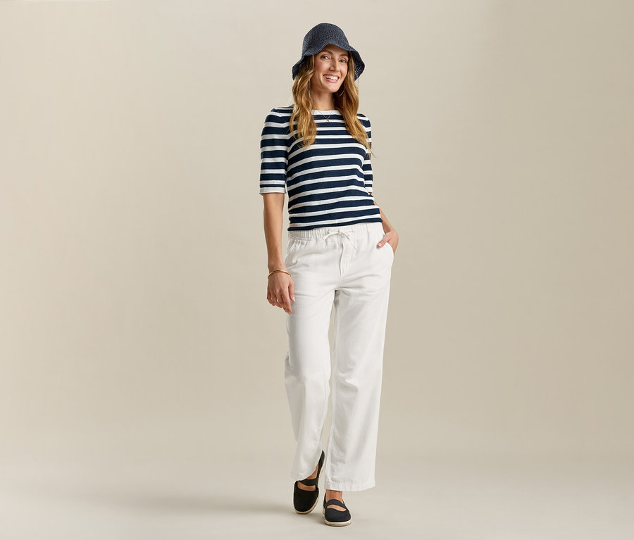 Frau mit blau gestreiftem Shirt, Hut und Wide-Komfort-Jeans. Sie steht mit einer Hand in der Hosentasche vor hellem Hintergrund.