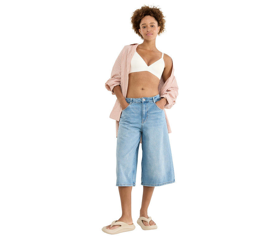 Frau steht mit Händen in den Hosentaschen, trägt ein weißes Bralette, eine rosa gestreifte Bluse, hellblaue Culottes und beigefarbene Zehentrenner.