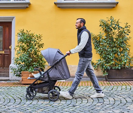 Mann schiebt einen grauen Kinderwagen auf einem gepflasterten Weg vor einem gelben Gebäude.