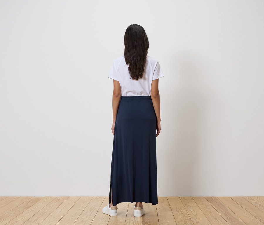 Rückansicht einer Frau mit dunklen Haaren, die ein weißes T-Shirt, einen langen, marineblauen Rock mit Schlitz und weiße Turnschuhe trägt.