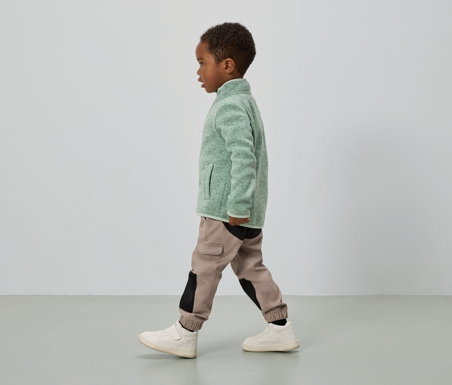 Ein Junge mit dunkler Haut geht, trägt eine grüne Fleecejacke, Cargo-Hosen mit schwarzen Einsätzen und cremefarbene Turnschuhe.