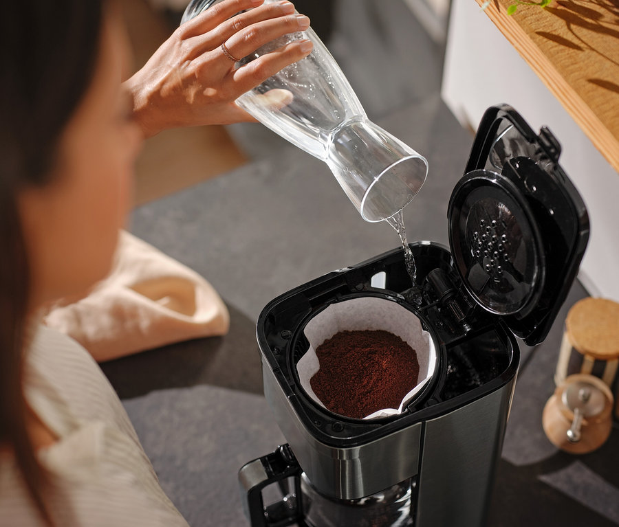 Frau gießt Wasser aus einer Glaskaraffe in eine Kaffeemaschine.