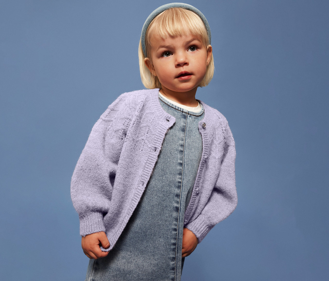 Mädchen mit blonden Haaren trägt eine helllila NAME IT Kinder-Strickjacke über einem Jeanskleid vor blauem Hintergrund.