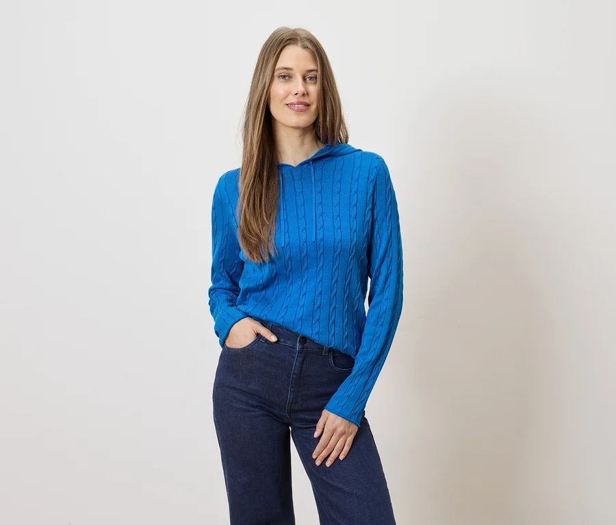 Frau mit langen Haaren posiert in blauer Strickjacke mit Kapuze und dunkler Jeans vor hellem Hintergrund.