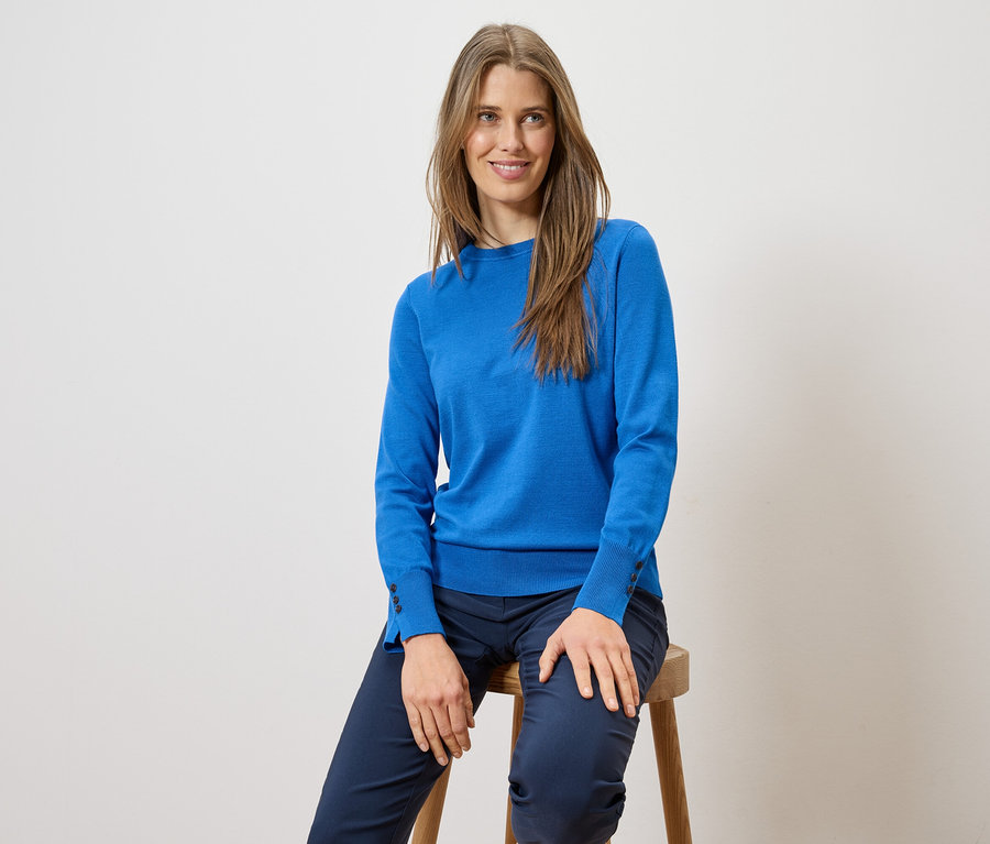 Frau mit langen Haaren sitzt auf einem Hocker und trägt einen blauen Pullover und eine blaue Hose.