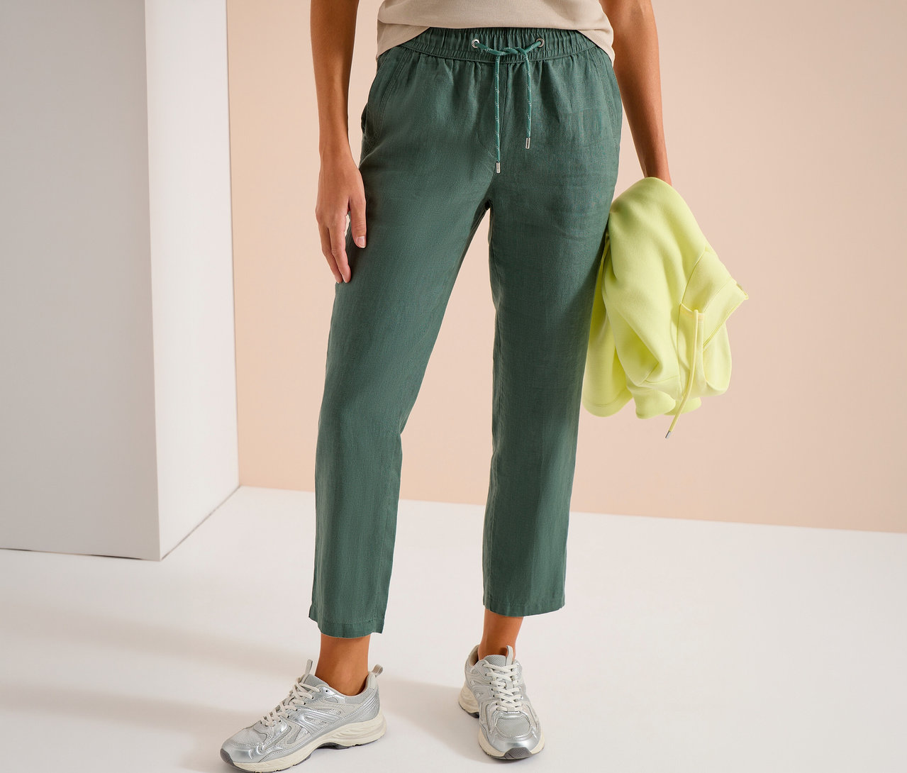 Frau trägt grüne Hose mit Kordelzug und silberne Turnschuhe, hält gelben Hoodie.