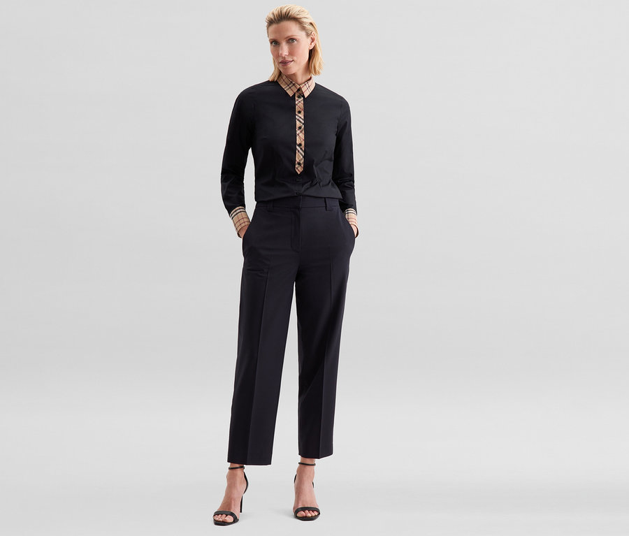 Eine Frau trägt eine schwarze Seidensticker Popeline-Bluse mit Karodetail an Kragen und Manschetten, dazu eine schwarze Hose und schwarze Sandalen mit hohen Absätzen.