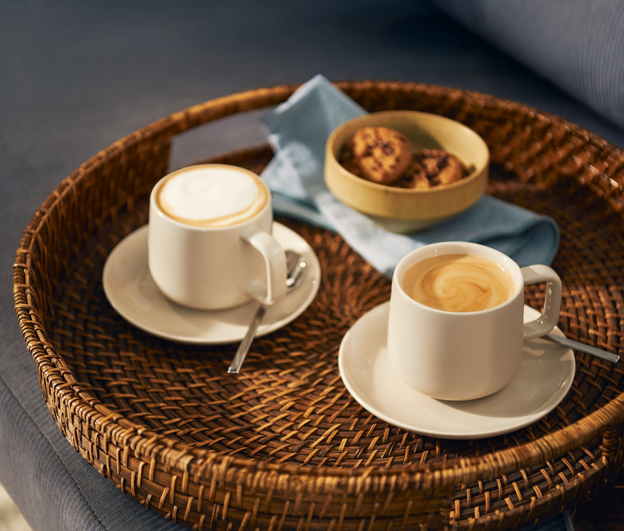 Zwei helle Kaffeetassen, eine mit Milchschaum und eine mit Latte-Art-Wirbel, stehen auf passenden Untertassen mit Löffeln auf einem runden geflochtenen Tablett. Eine kleine Schale mit Keksen und eine hellblaue Serviette sind ebenfalls auf dem Tablett.