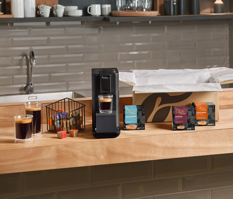 Schwarze Qbo Iconic, Dark Chrome 1,1 DAEE Kaffeemaschine auf einer Holztheke, mit einem Glas Kaffee darunter. Daneben stehen zwei weitere Kaffeegläser, ein Korb voller Qbo Kapseln und lose Kapseln. Rechts sind offene Qbo Caffè Grande KIRINYAGA BAYA, Qbo Espresso APANECA ANA und Qbo Caffè MINAS VERDE Kapselpackungen zu sehen.