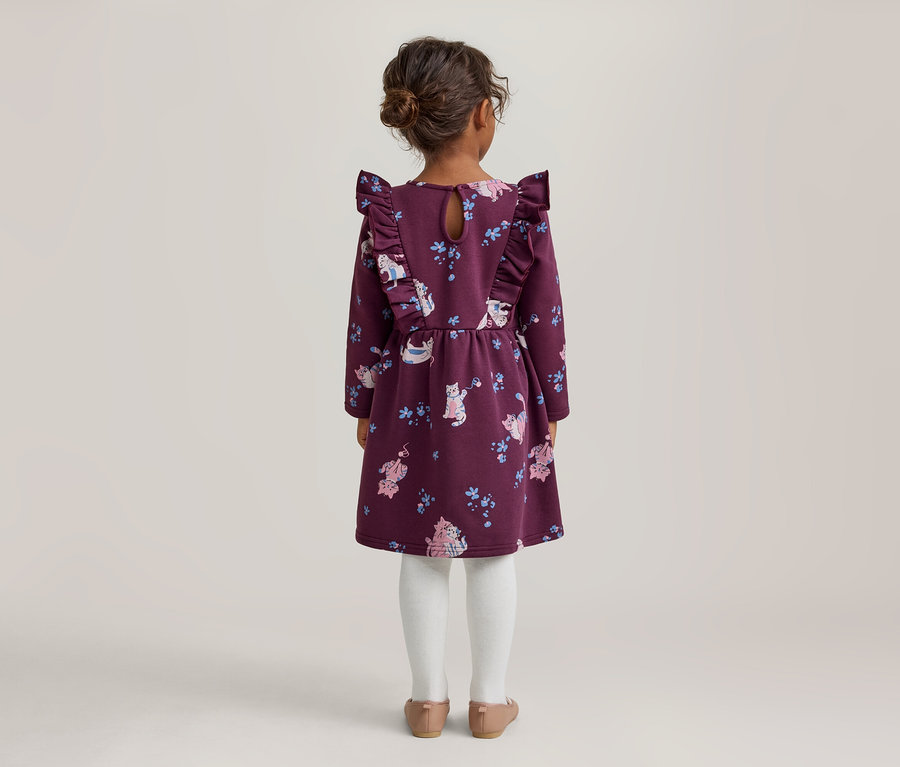 Kleines Mädchen von hinten in einem lila Kleid mit Katzen- und Blumenmuster, Rüschen an den Schultern, weißer Strumpfhose und hellbraunen Schuhen.