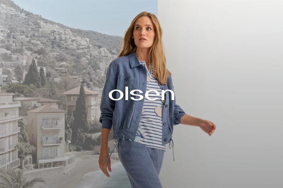 Frau in blauer Jacke, gestreiftem Oberteil und blauer Hose, die vor einem geteilten Hintergrund aus Küstenstadt und grauer Wand geht, mit dem Schriftzug 'olsen'.