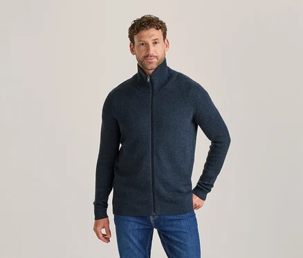 Mann in dunkelblauer Strickjacke mit Reißverschluss und blauen Jeans, steht und blickt in die Kamera.
