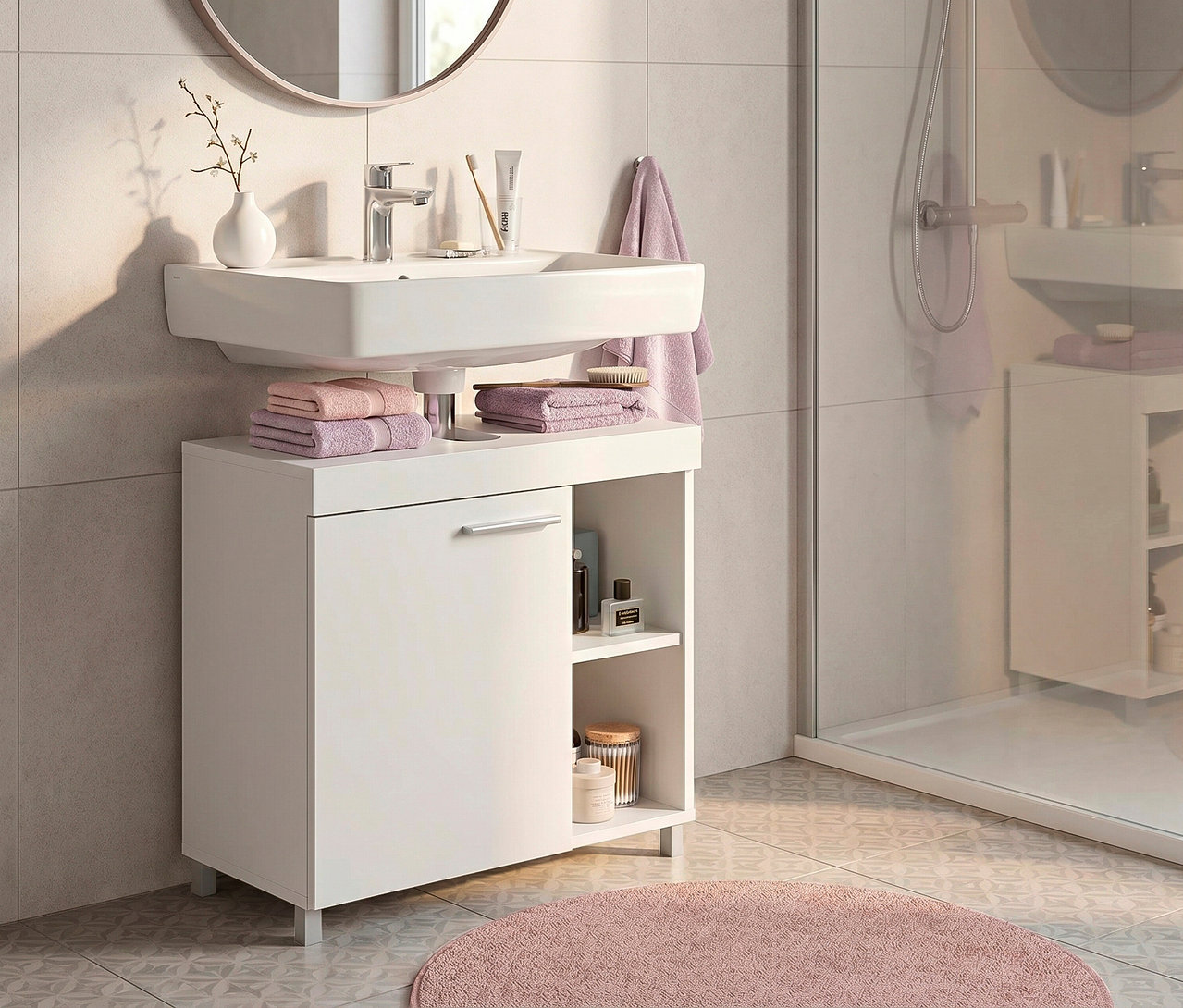 Badezimmer mit weißem Waschbeckenunterschrank und rechteckigem Waschbecken, darüber ein runder Spiegel. Auf dem Schrank liegen gefaltete rosa und lila Handtücher. Rechts daneben ist eine Duschkabine zu sehen, davor liegt ein runder rosa Badteppich.