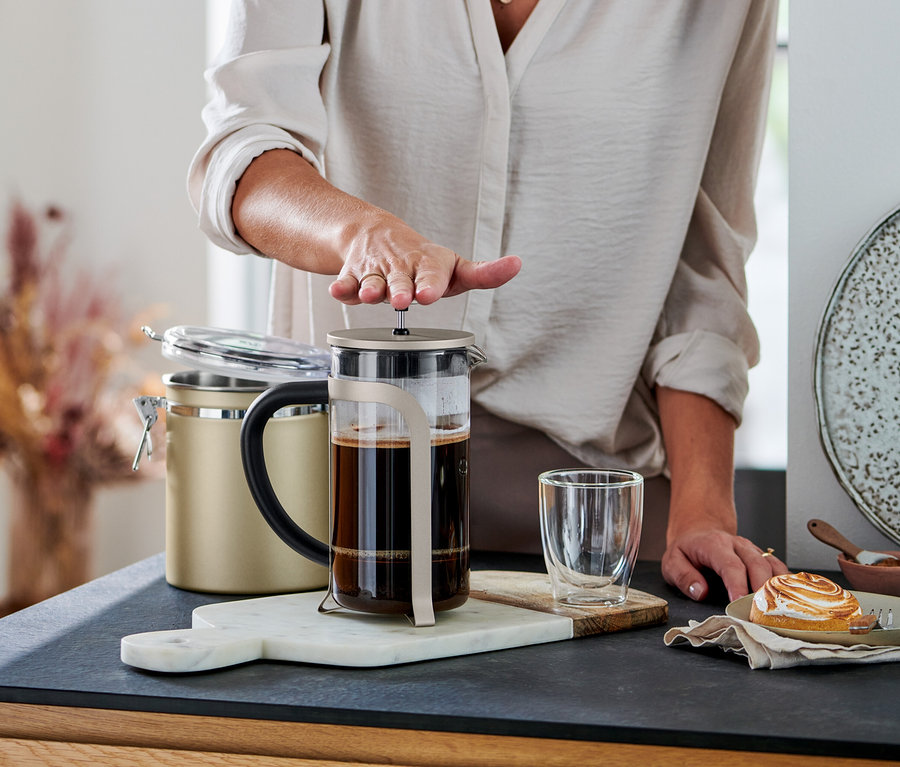 Eine Person drückt den Stempel einer champagnerfarbenen Siebstempelkanne (800 ml, 6 Tassen) mit Kaffee herunter. Daneben stehen ein Glas und ein Gebäck.