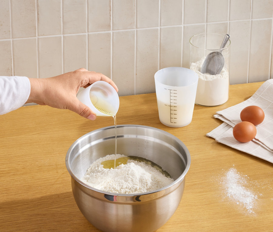 Eine Hand gießt Flüssigkeit aus einem Messbecher in eine Schüssel mit Mehl und einem aufgeschlagenen Ei. Auf der Arbeitsplatte stehen ein weiterer Messbecher, ein Mehlglas, zwei Eier auf einem Tuch und verschüttetes Mehl.