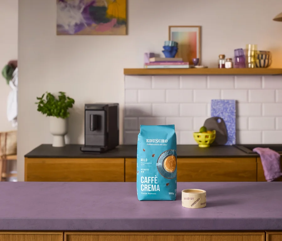 Eine Packung Eduscho Caffè Crema Mild Kaffeebohnen und ein kleiner Eduscho Behälter stehen auf einer violetten Arbeitsplatte. Im Hintergrund ist ein schwarzer «Esperto mini» Kaffeevollautomat in einer Küche zu sehen.