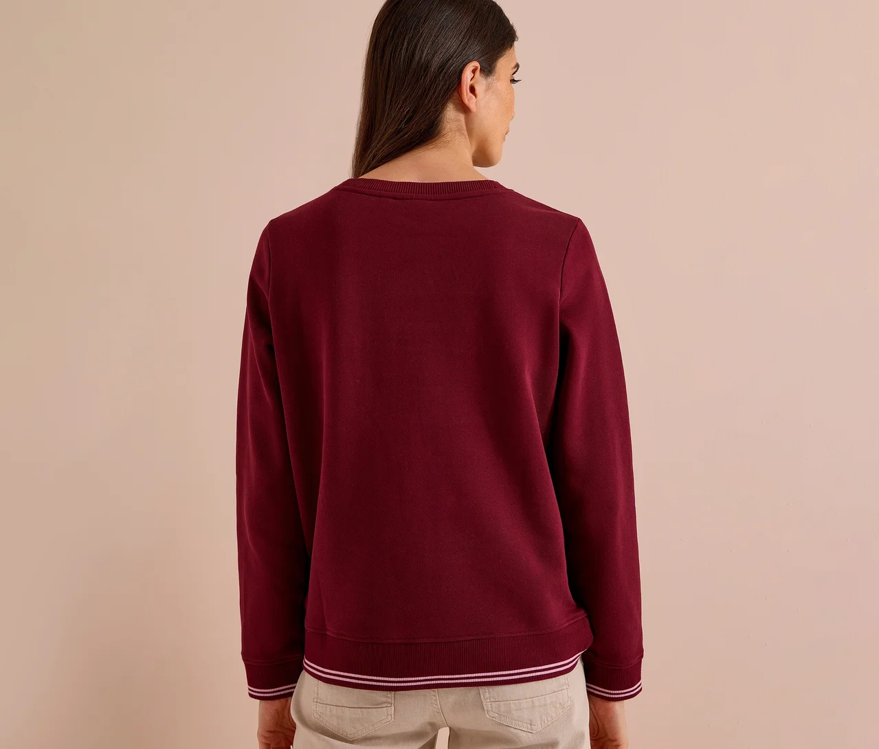 Frau von hinten in einem bordeauxroten CECIL Sweatshirt mit weißen Streifen an Bündchen und Saum, dazu helle Hose.