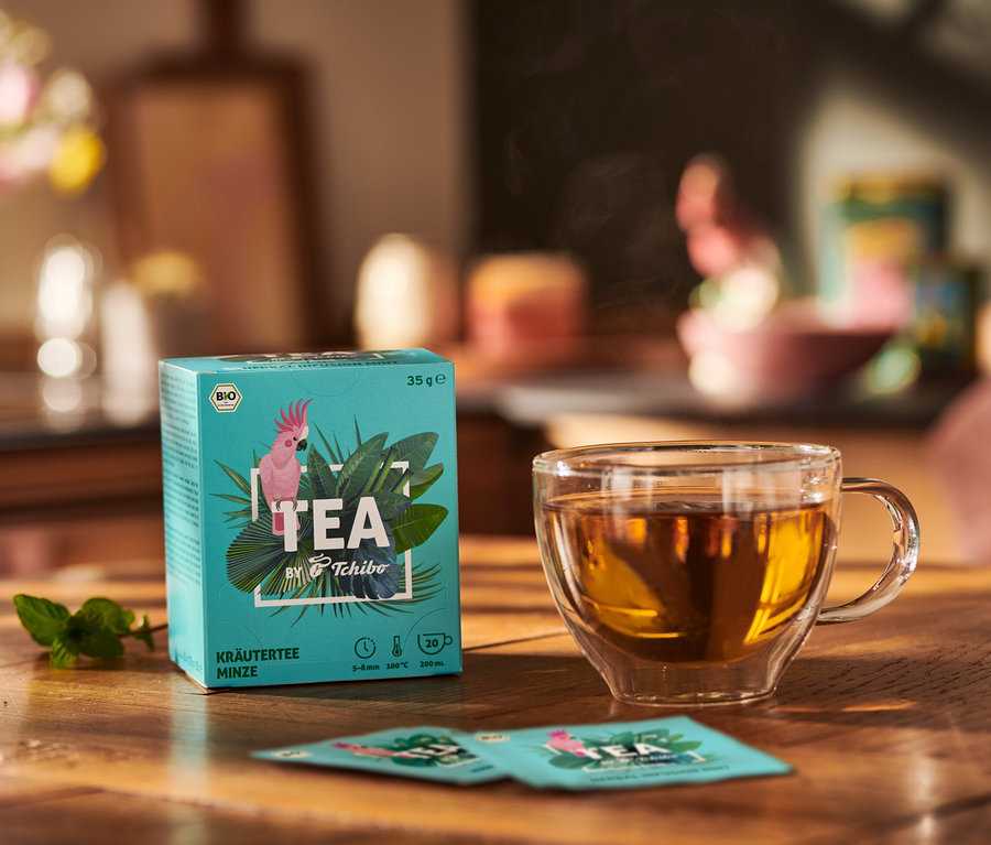 Packung TEA by Tchibo Bio Kräutertee Minze neben Teetasse und Minze Blätter auf Holztisch.