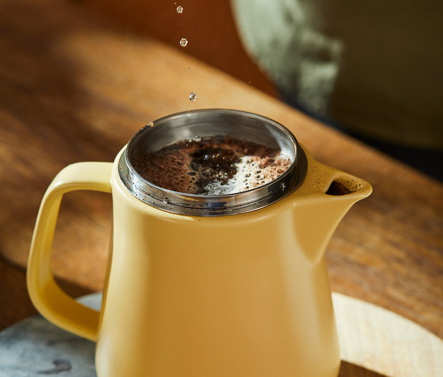 Wasser wird aus einem Wasserkocher in einen gelben Kaffeebehälter auf einem Holztisch gegossen.