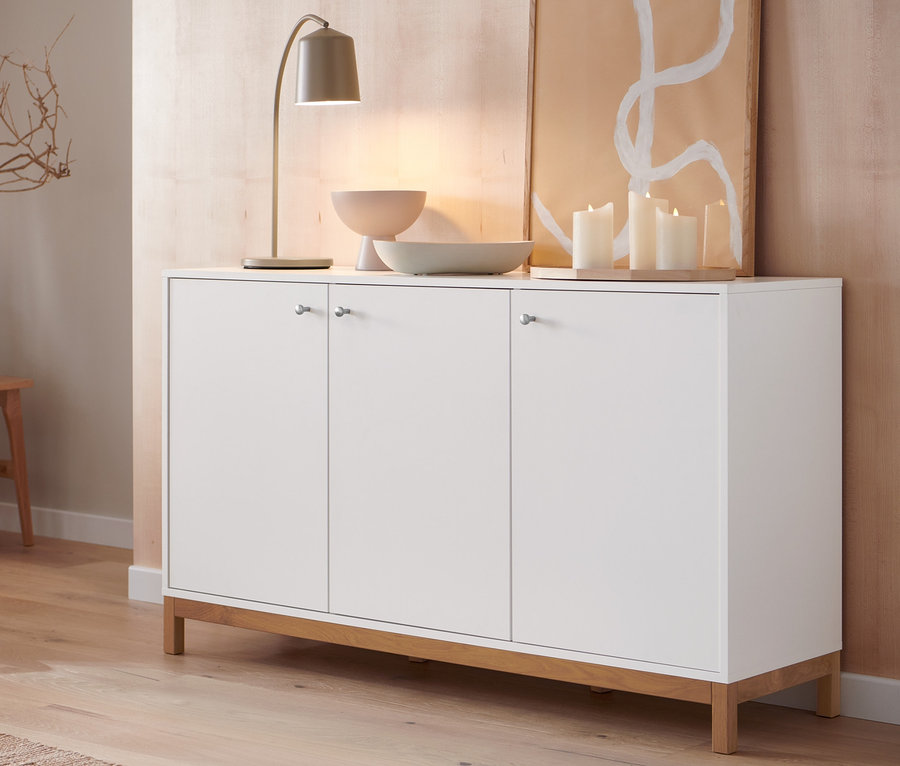 Eine creme Tischleuchte aus Metall steht auf einem weißen Sideboard neben einer Schale und Kerzen.