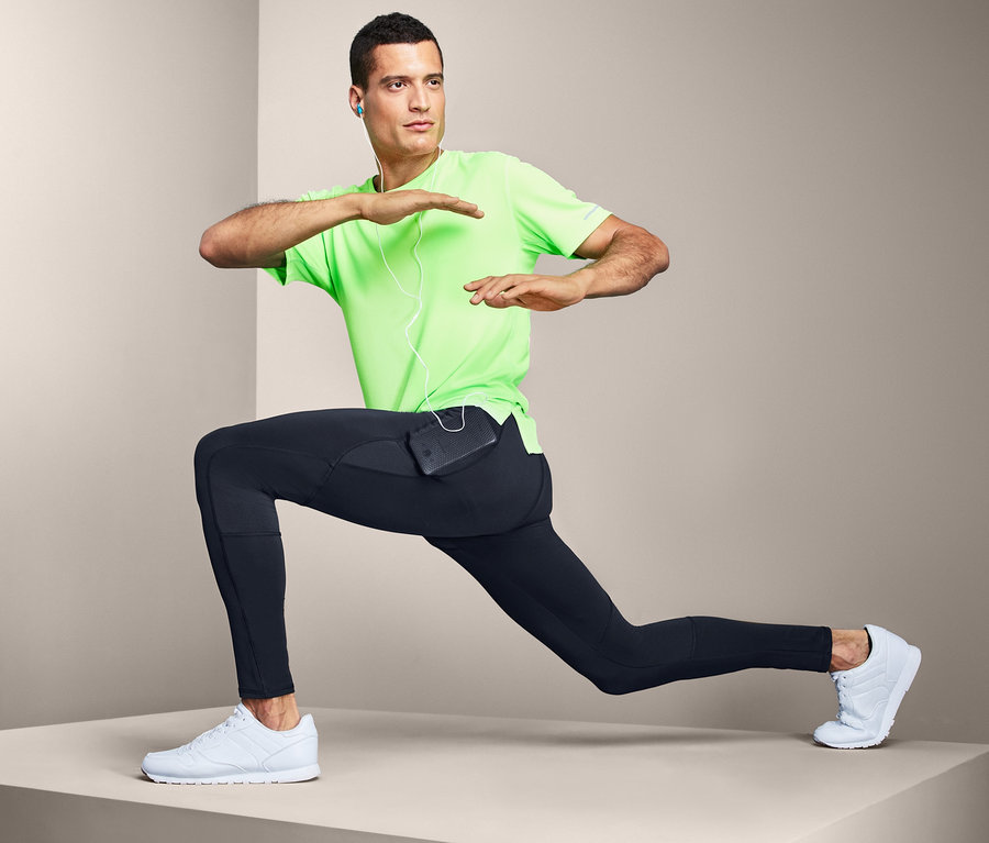 Mann in grünem T-Shirt und Sporttight macht Ausfallschritte. In der Tasche der Tights befindet sich ein Mobiltelefon.