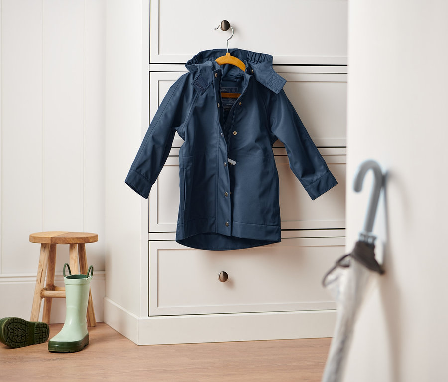 Eine dunkelblaue Regenjacke hängt an einer weißen Kommode. Ein Holzhocker und ein Paar grüne Gummistiefel stehen auf dem Boden.