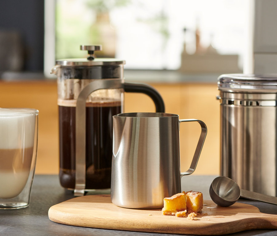 Ein Glas Kaffee steht neben einer French Press und einem kleinen Milchkännchen aus Edelstahl auf einem Holzbrett.
