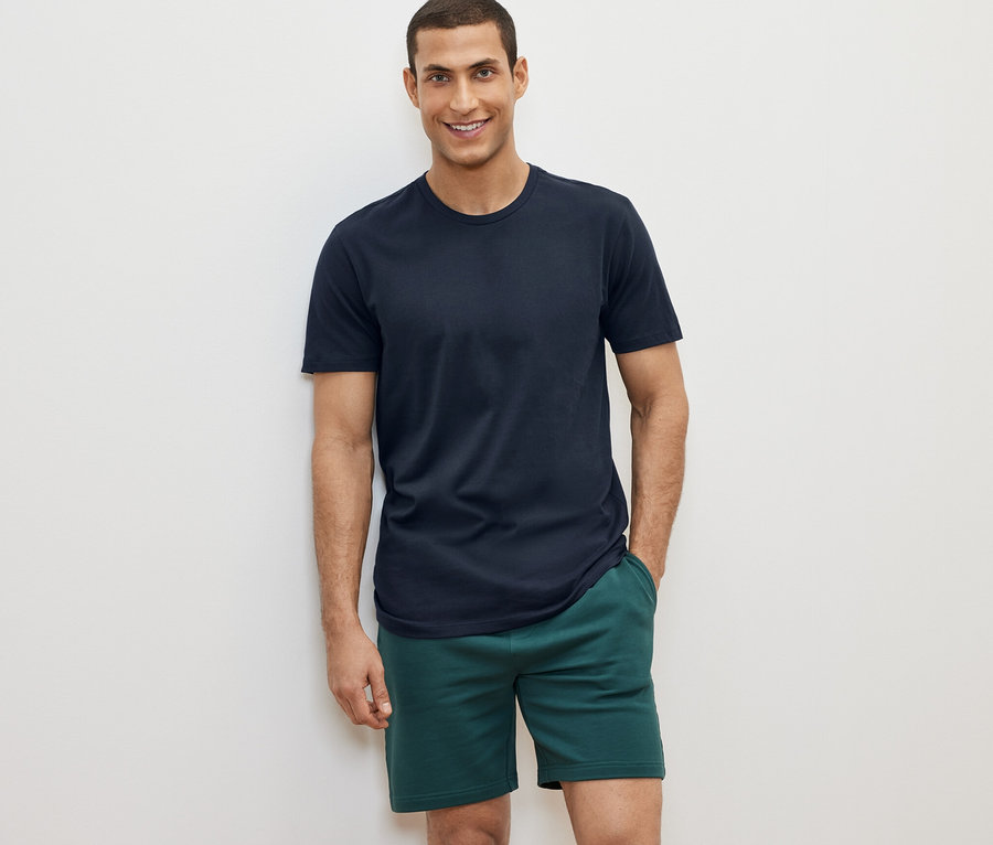 Mann posiert vor weißem Hintergrund in einem von 2 T-Shirts mit Rundhalsausschnitt, grüne Shorts.