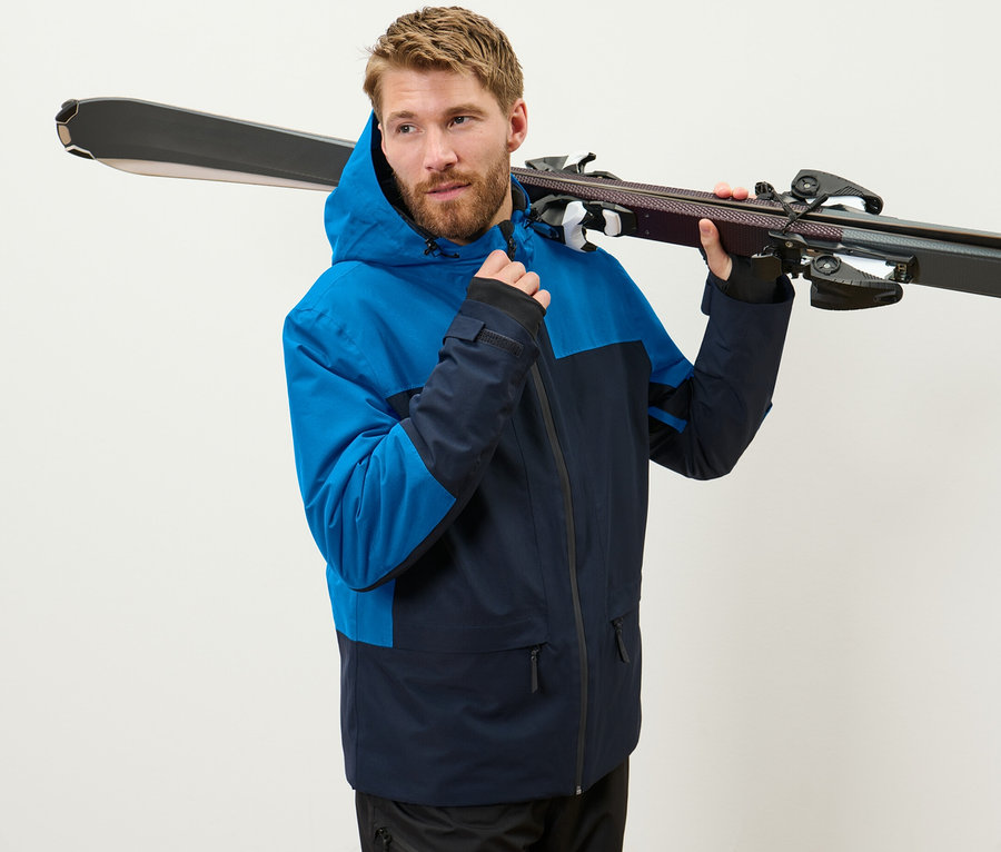 Mann in blauer Skijacke trägt Ski über der Schulter.