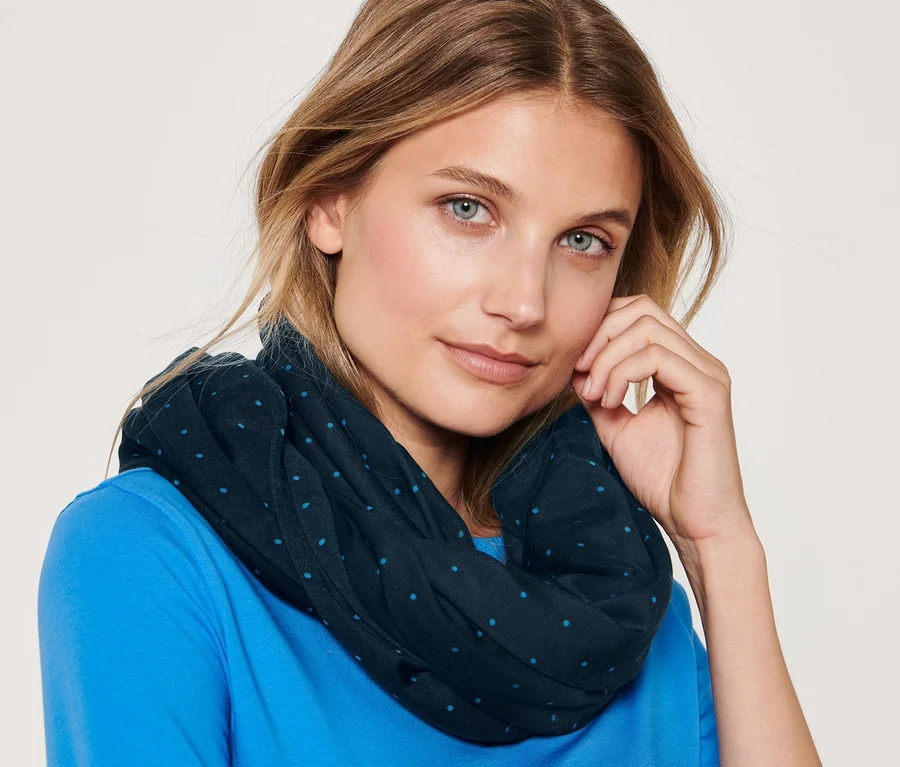 Porträt einer blonden Frau mit blauen Augen, die einen gepunkteten Loop-Schal und ein blaues T-Shirt trägt.