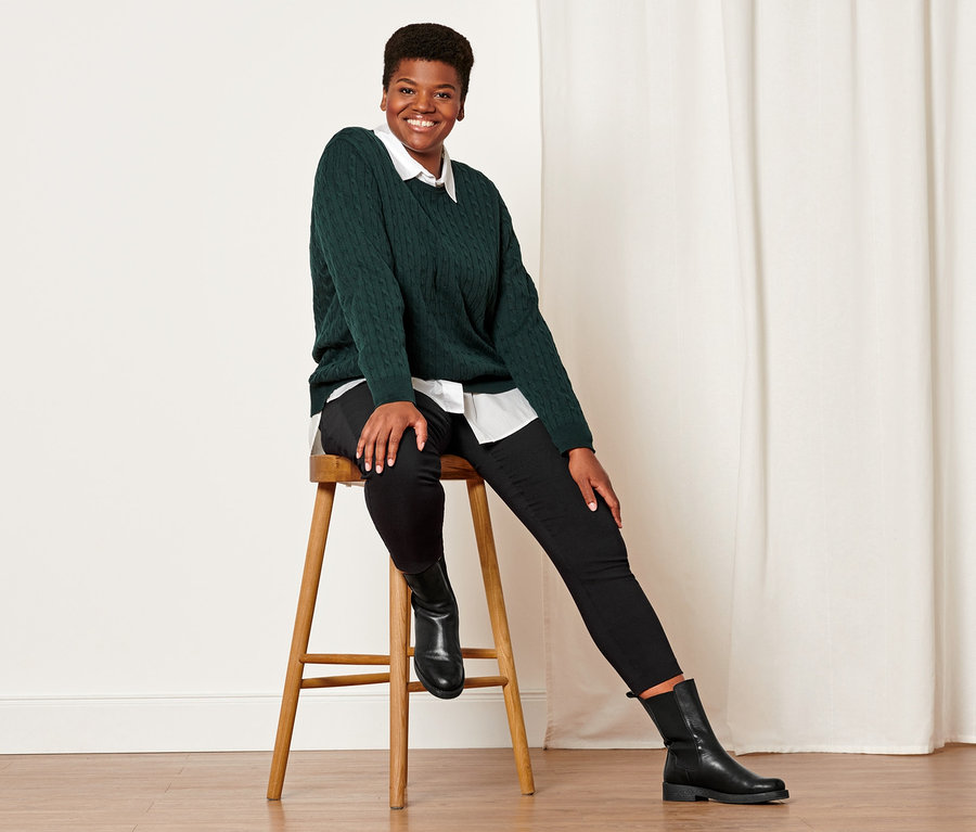 Frau sitzt auf einem Hocker und trägt einen dunkelgrünen Zopfstrickpullover, eine schwarze Stretchhose und Lederboots.