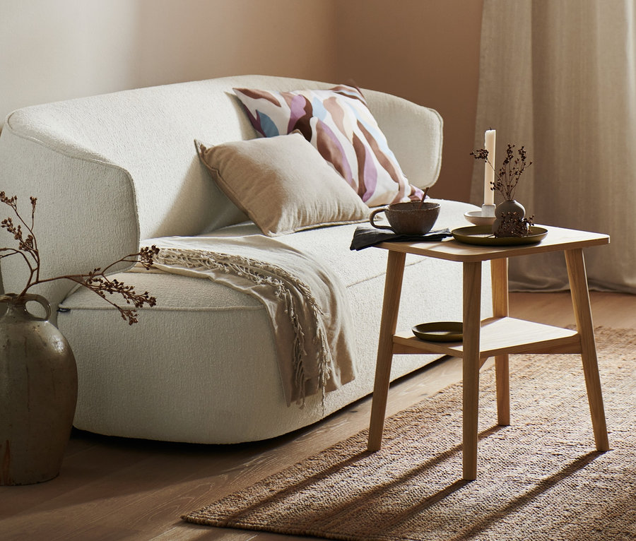 Ein Wohnzimmer mit einem beigen Sofa, auf dem eine Chenille-Wohndecke und Dekokissen liegen. Neben dem Sofa steht ein Beistelltisch mit 2 Dekotabletts. Im Raum hängen 2 Vorhangschals und an der Decke hängt eine Deckenleuchte mit Filzschirm.