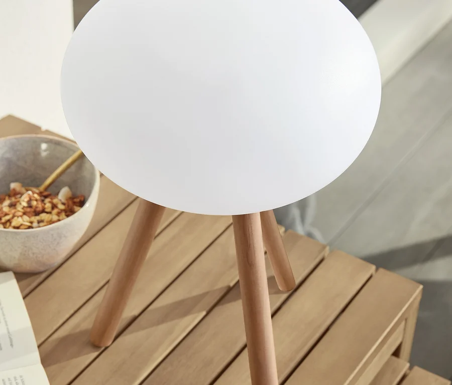 Auf einem Holztisch stehen eine LED-Akku-Tischleuchte mit Beinen aus Massivholz, eine Schale mit Essen und ein offenes Buch.