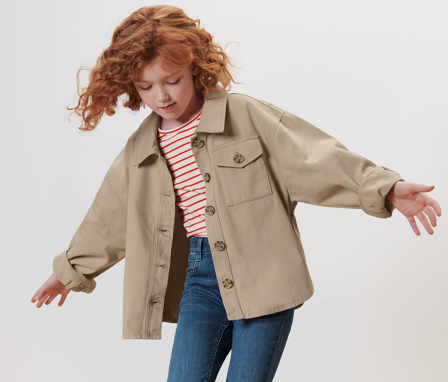 Mädchen mit lockigen roten Haaren winkt mit den Armen und trägt eine Kinder-Denimjacke.